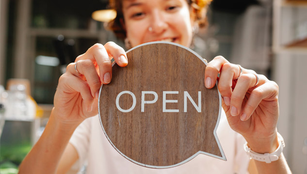 A sign with the word "OPEN" shaped like a speech bubble is held up by a young woman wearing a white shirt and white watchband