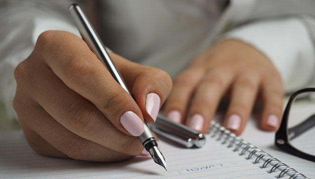A Latina woman with manicured light pink nails holds a pen over a notebook, preparing to write.